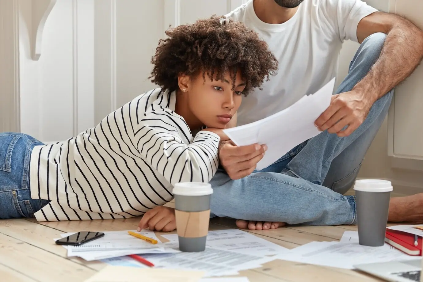 Una gerente y un gerente posan en el suelo, discuten un informe, aprenden los resultados de ventas a partir de papeles, se reúnen en un ambiente doméstico, beben café de vaso desechable y analizan los beneficios de la empresa. Concepto de colaboración.